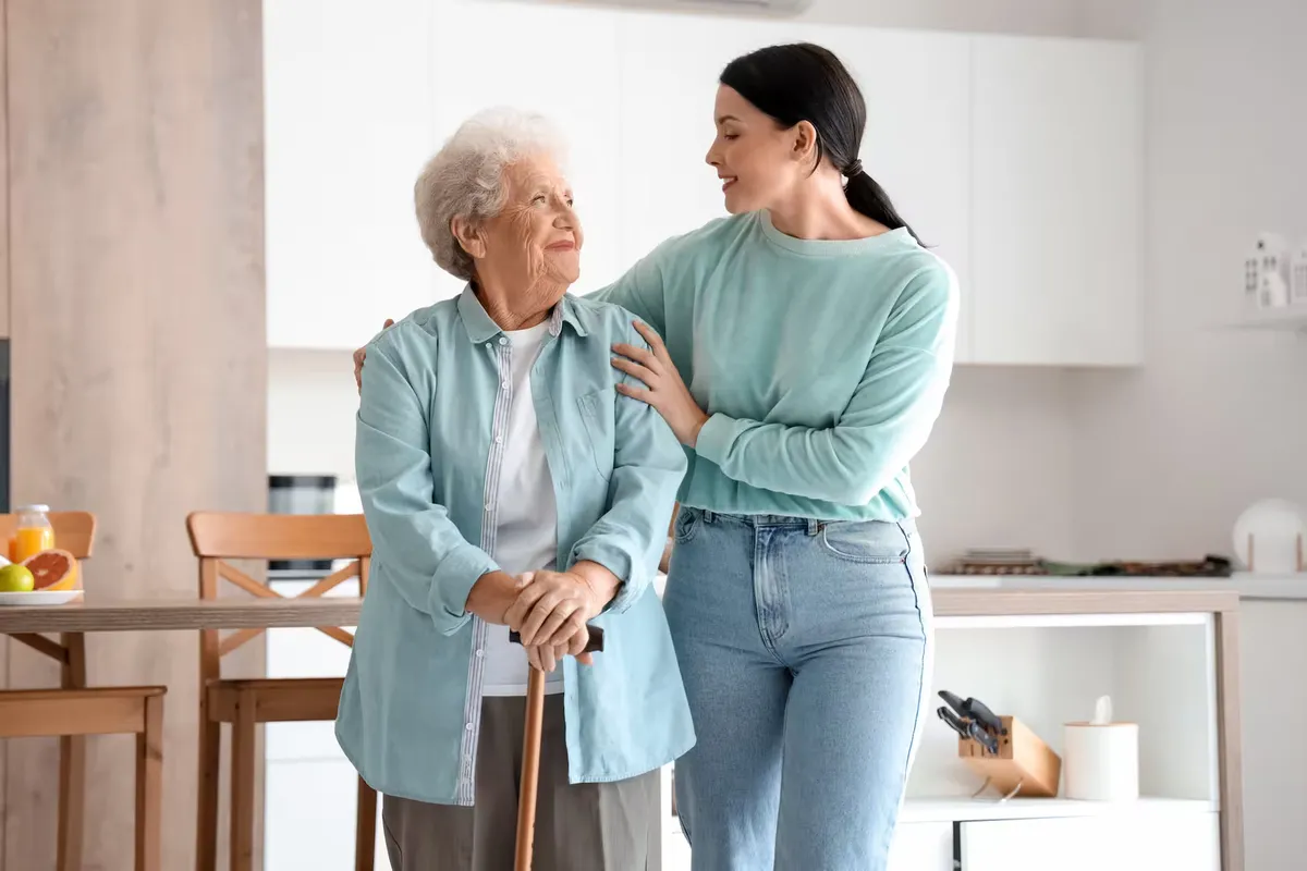 A carer supporting an elderly woman in a bright living room